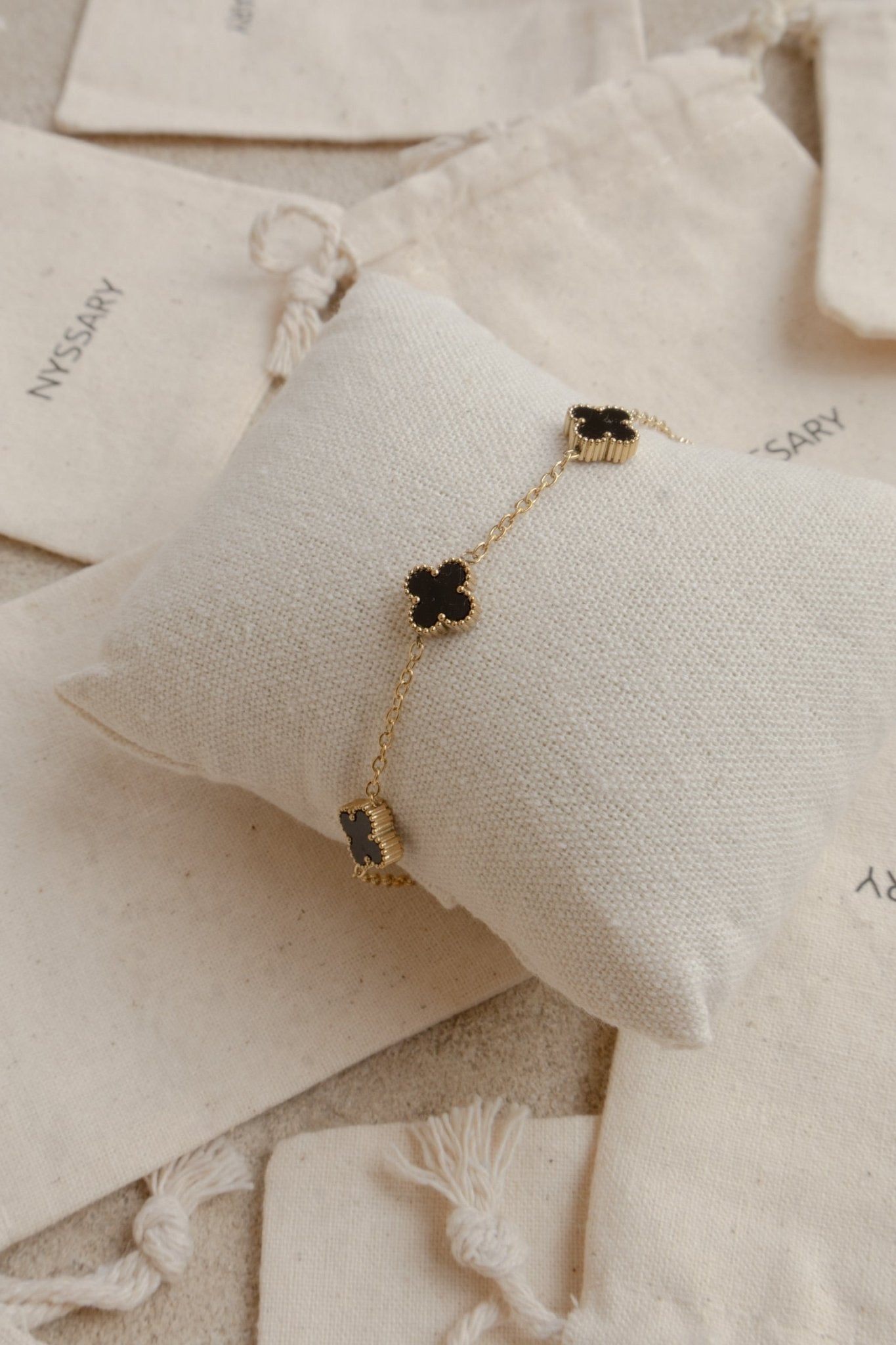 Bracelet en acier inoxydable doré avec trèfles noirs, présenté sur coussin bijou posé sur des pochettes Nyssary