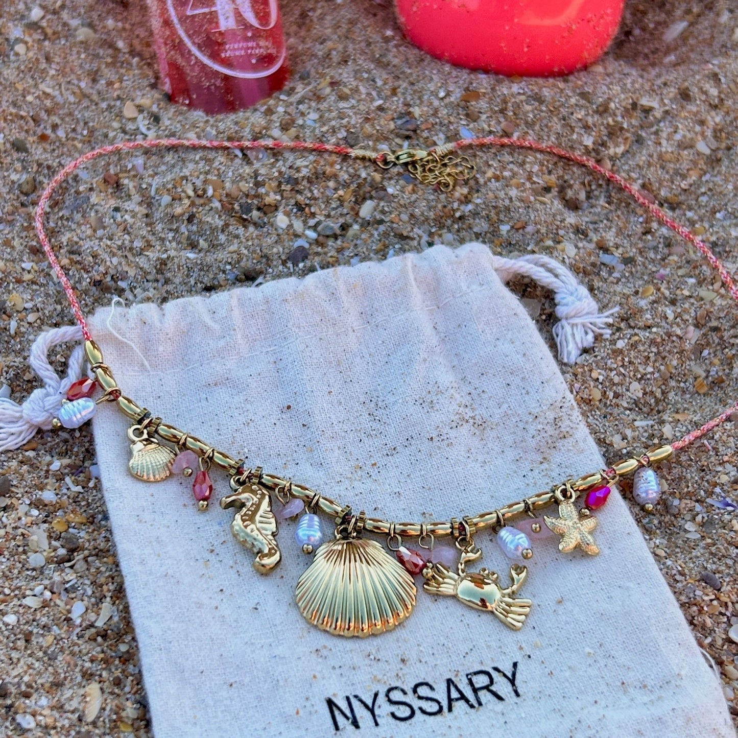 Collier doré Océane avec charms marins, présenté sur des pochettes Nyssary avec un fond sablé