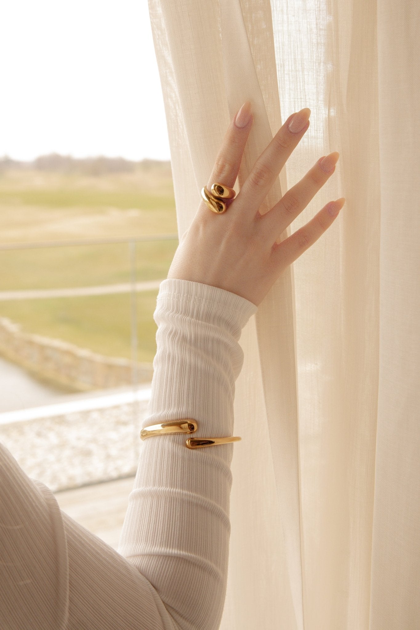 Femme près de la fenêtre tenant un rideau, portant au poignet un bracelet et une bague acier inoxydable dorée au doigt