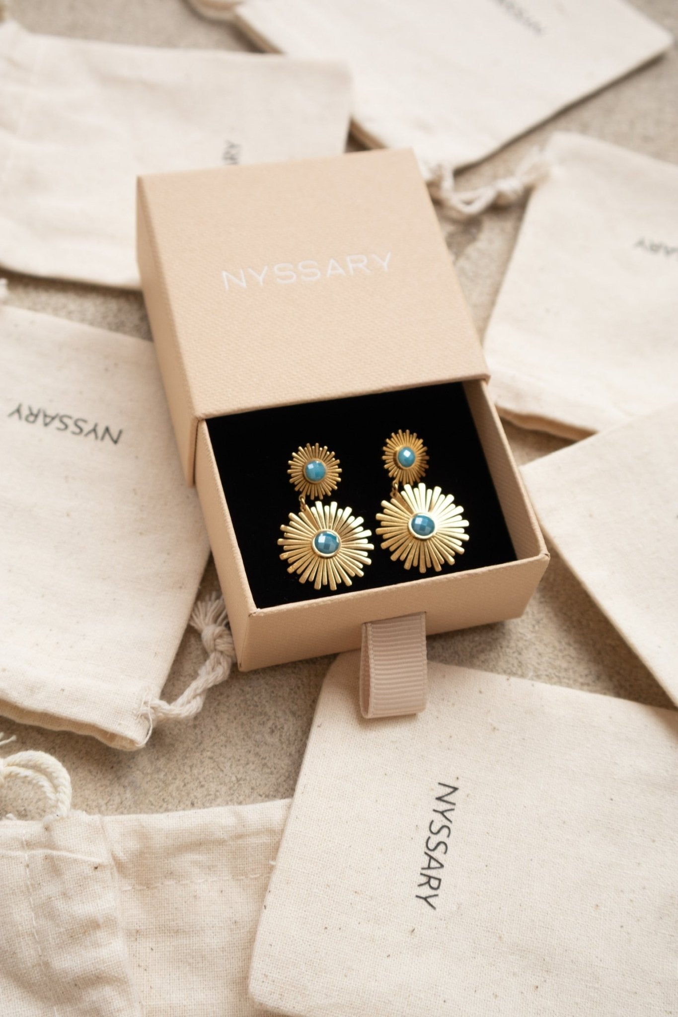 Boucles oreilles dorées Daisy présentées sur des pochettes Nyssary