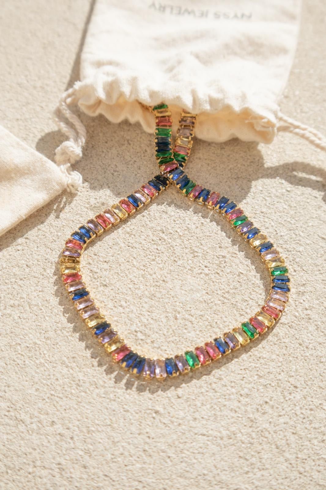 Collier acier inoxydable Arcadia avec strass multicolores, présenté sur des pochettes Nyssary avec un fond sablé