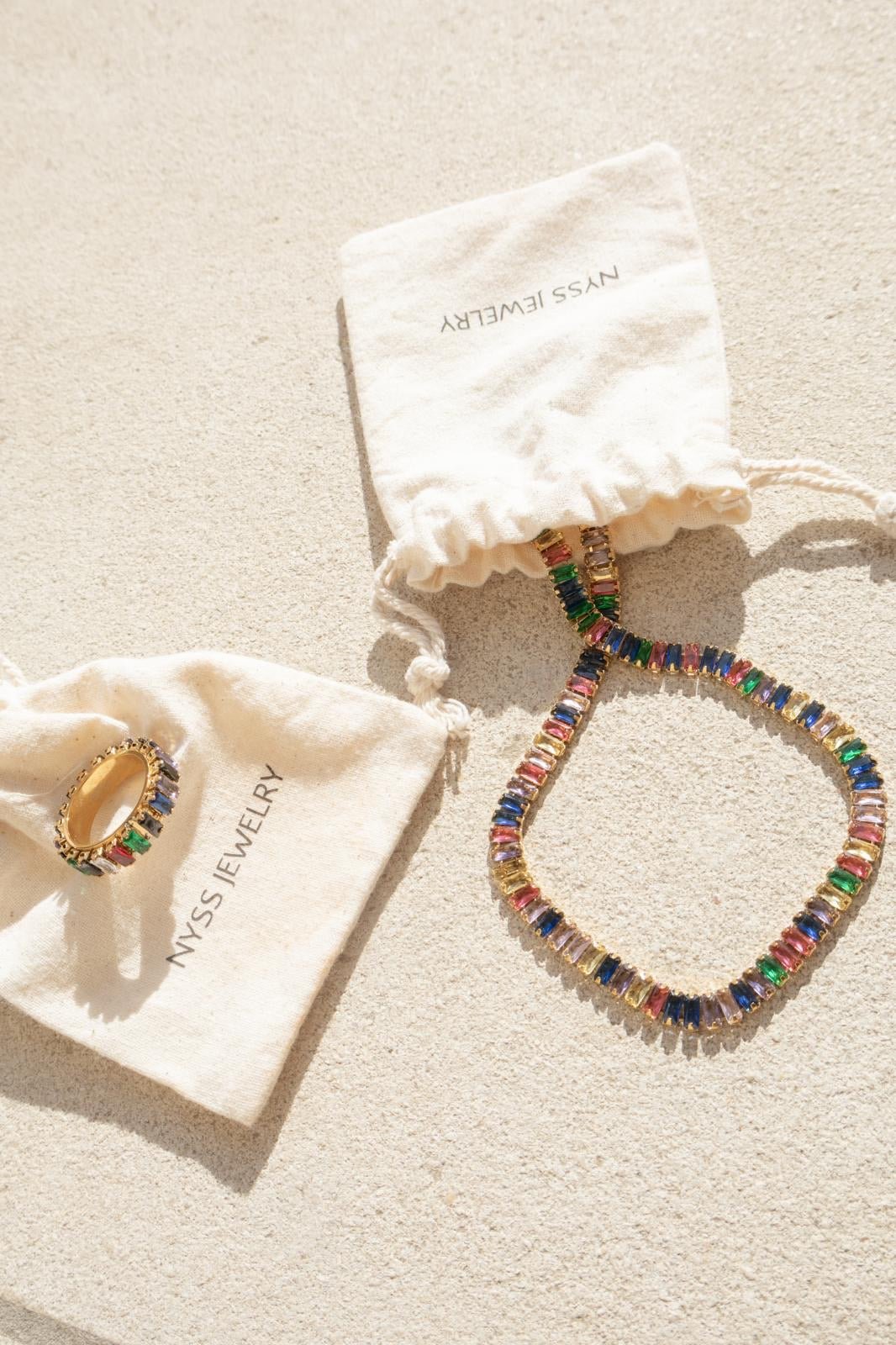 Collier acier inoxydable Arcadia avec strass multicolores, présenté sur des pochettes Nyssary avec un fond sablé