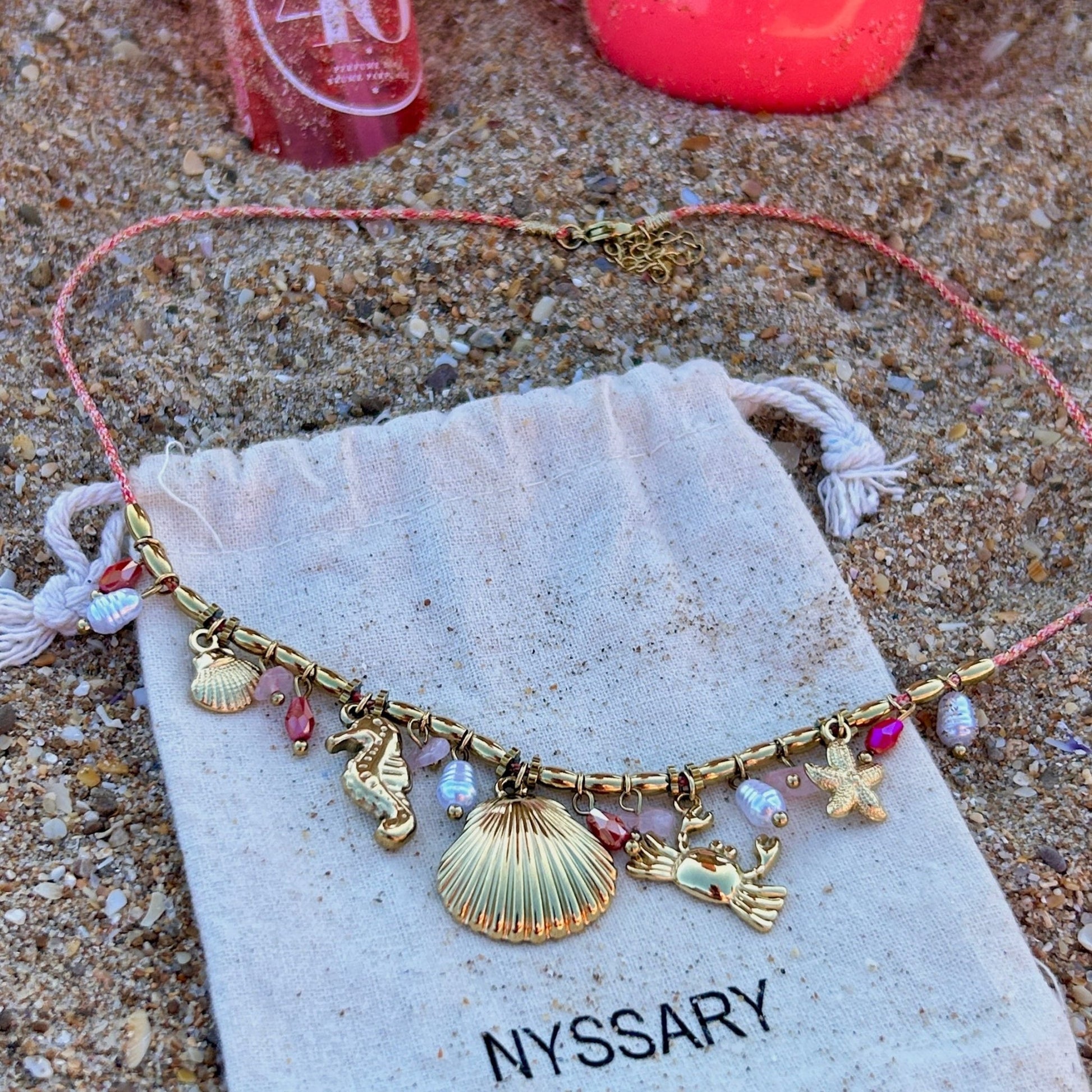Collier doré Océane avec charms marins, présenté sur des pochettes Nyssary avec un fond sablé