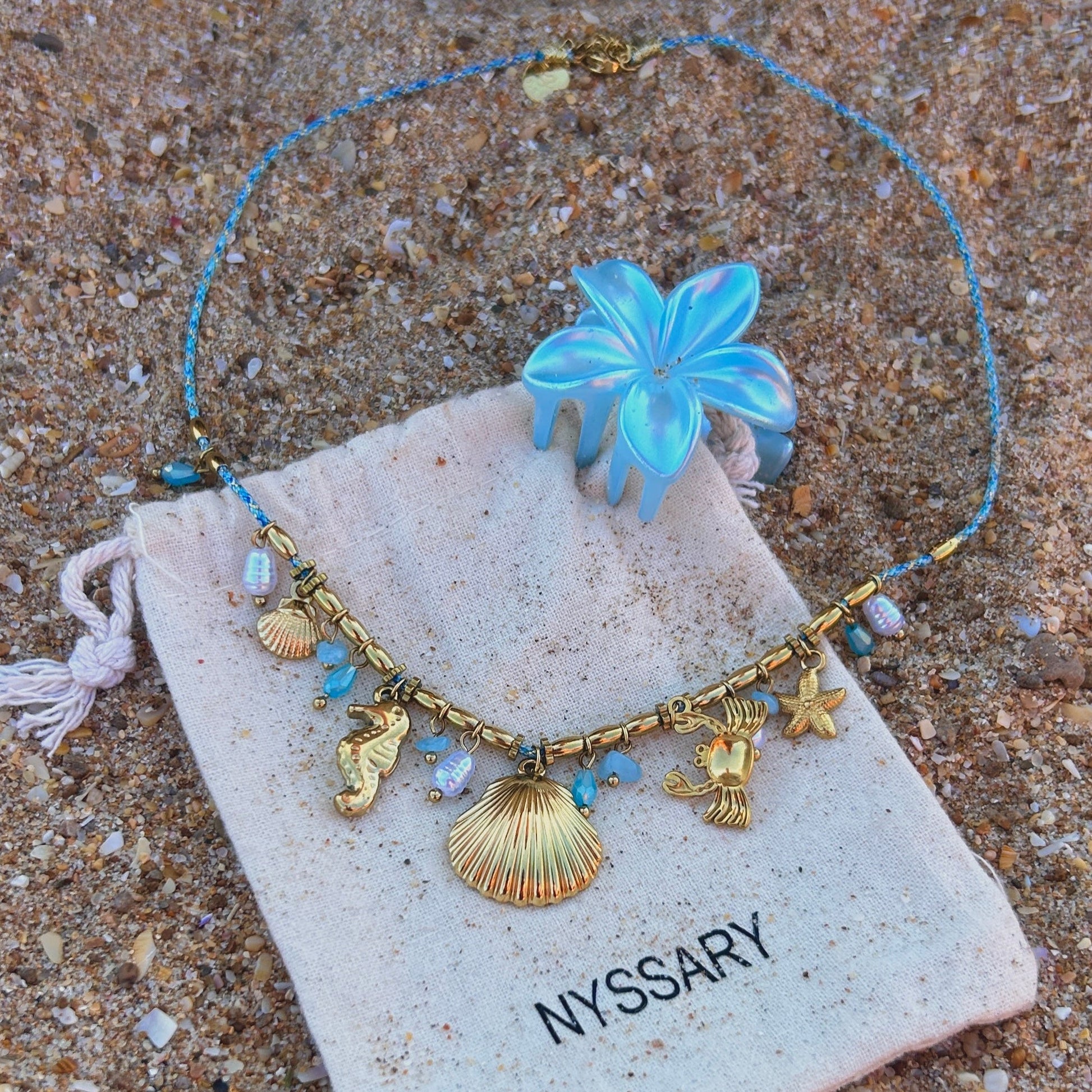 Collier doré Océane avec charms marins, présenté sur des pochettes Nyssary avec un fond sablé
