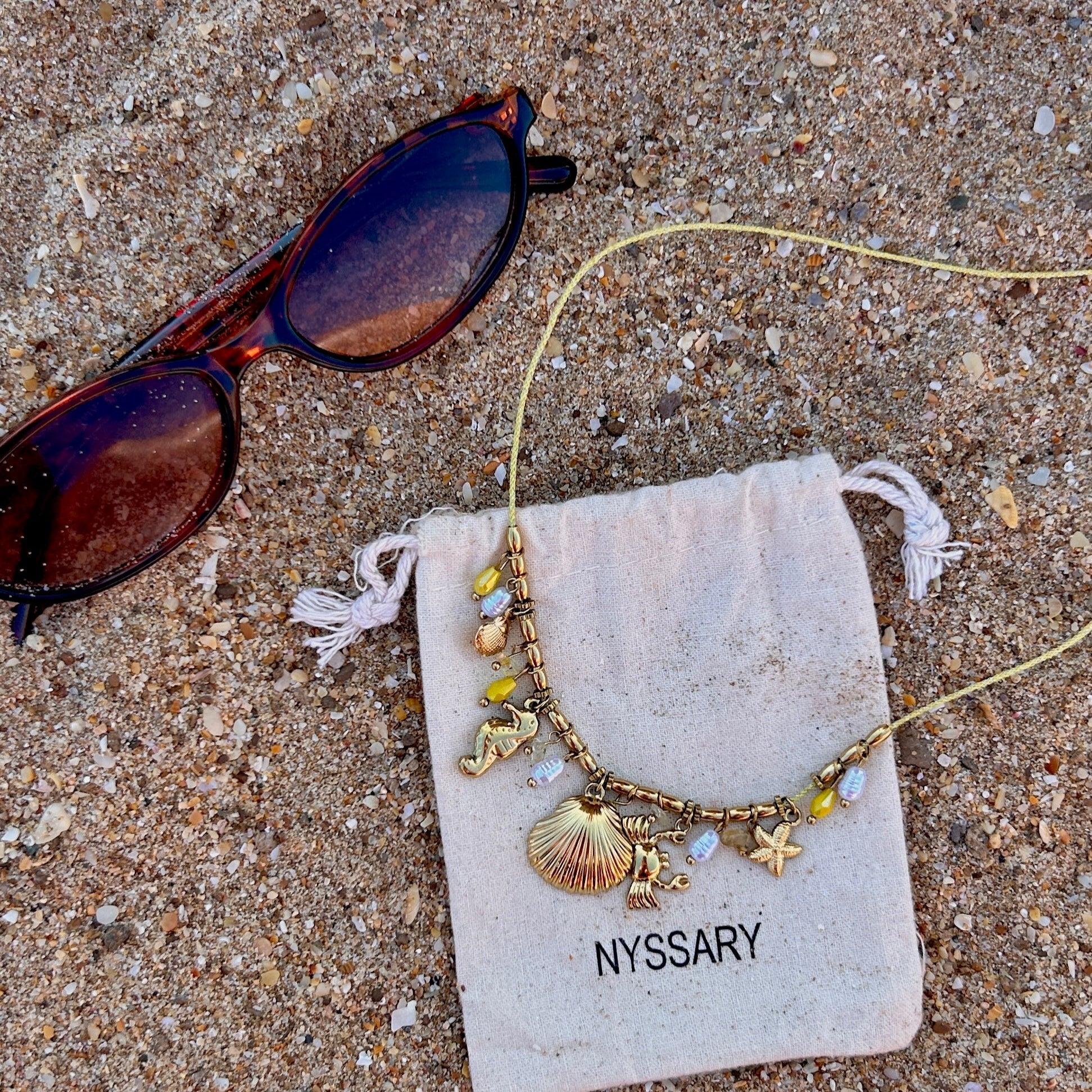 Collier doré Océane avec charms marins, présenté sur des pochettes Nyssary avec un fond sablé