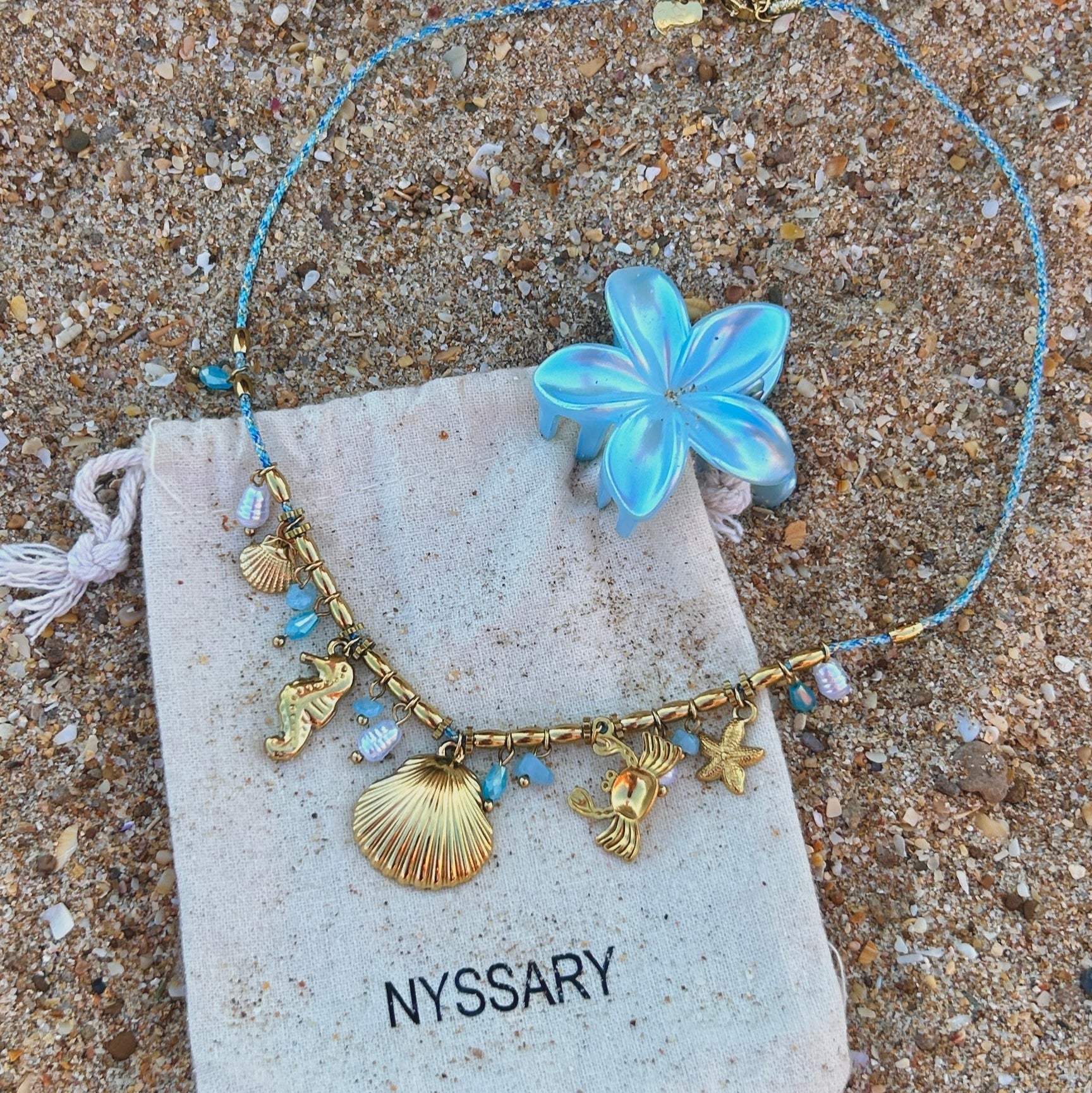 Collier doré Océane avec charms marins, présenté sur des pochettes Nyssary avec un fond sablé