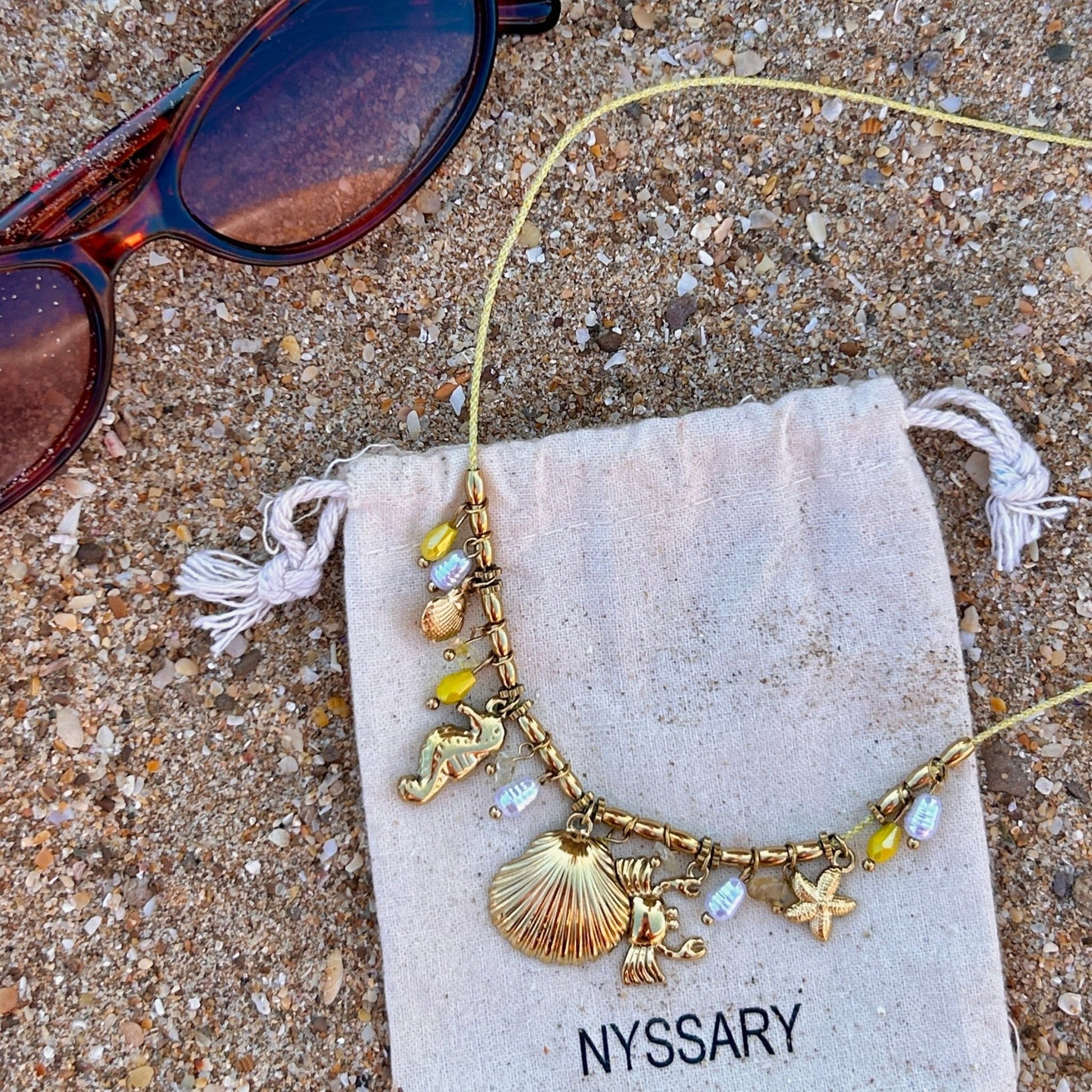 Collier doré Océane avec charms marins, présenté sur des pochettes Nyssary avec un fond sablé