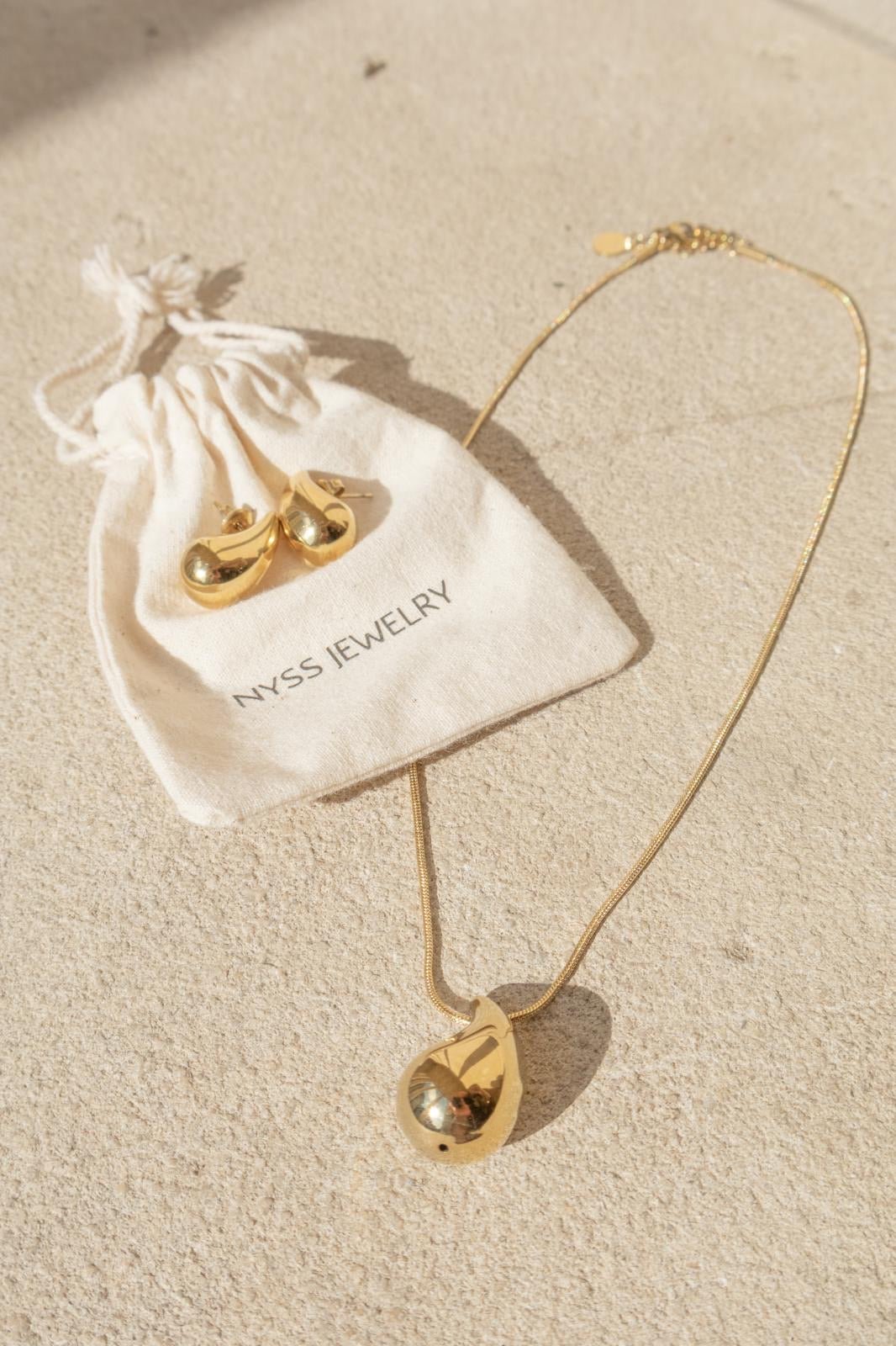 collier doré avec pendentif goutte Romy sur fond sablé, posé sur une pochette Nyssary